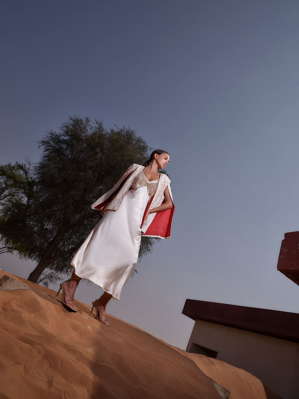 Woman in a white dress with a cape from the signature collection of Kashesh Global standing on a sand dune against a clear blue sky | Luxury Sustainable Women's Fashion in Dubai
