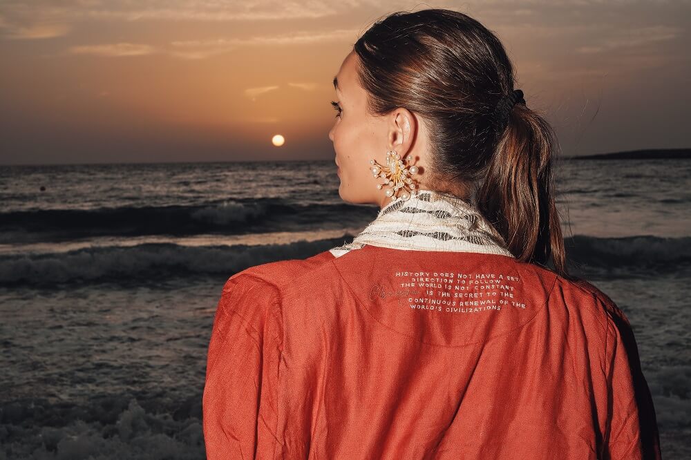 Woman in a coat from the signature collection of Kashesh Global looking at the sunset over the ocean | Luxury Sustainable Women's Fashion in Dubai